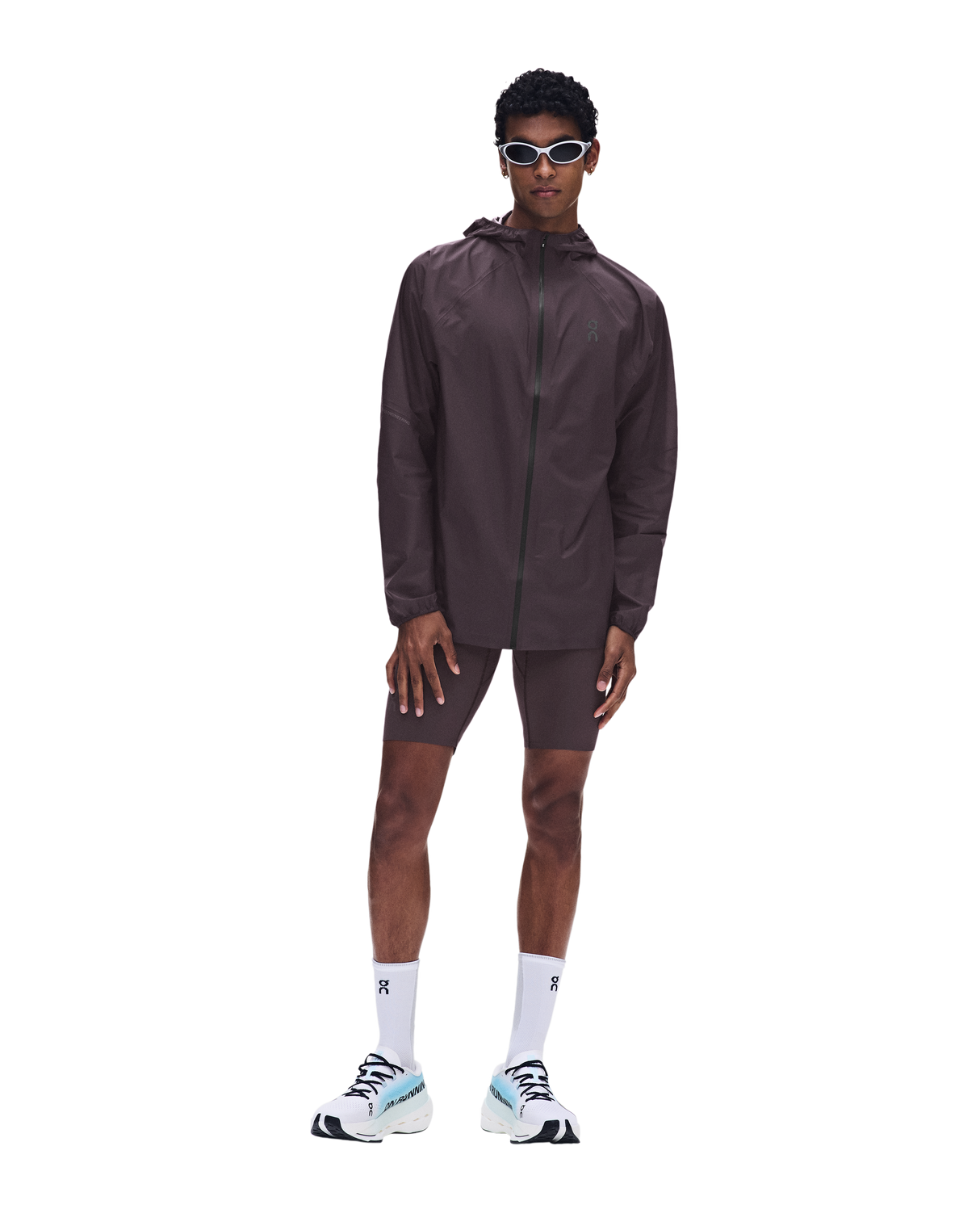 A male model wearing a dark brown athletic windbreaker jacket, matching shorts, white socks, and white sneakers with light blue accents, standing against a solid white background. - On Ultra Jacket (Men's) - Urchin