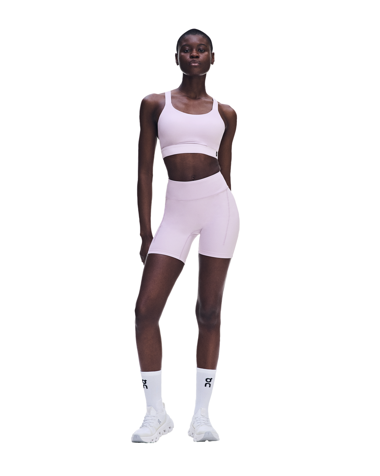 A model wearing a light pink athletic set consisting of a sports bra and matching high-waisted biker shorts, paired with white socks and white sneakers, set against a solid white background. - On Performance Bra (Women's) - Foxglove