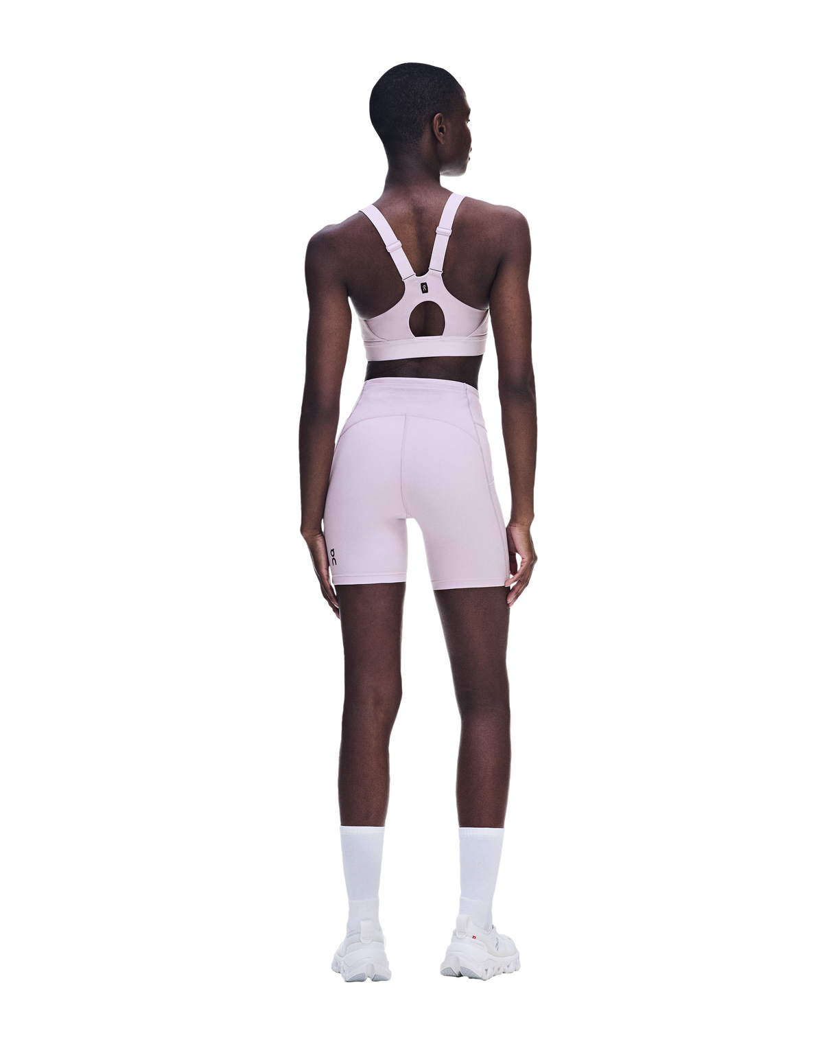 A model shown from the back wearing a light pink athletic set consisting of a sports bra with an open back and matching high-waisted shorts, paired with white socks and white sneakers on a solid white background. - On Performance Bra (Women's) - Foxglove