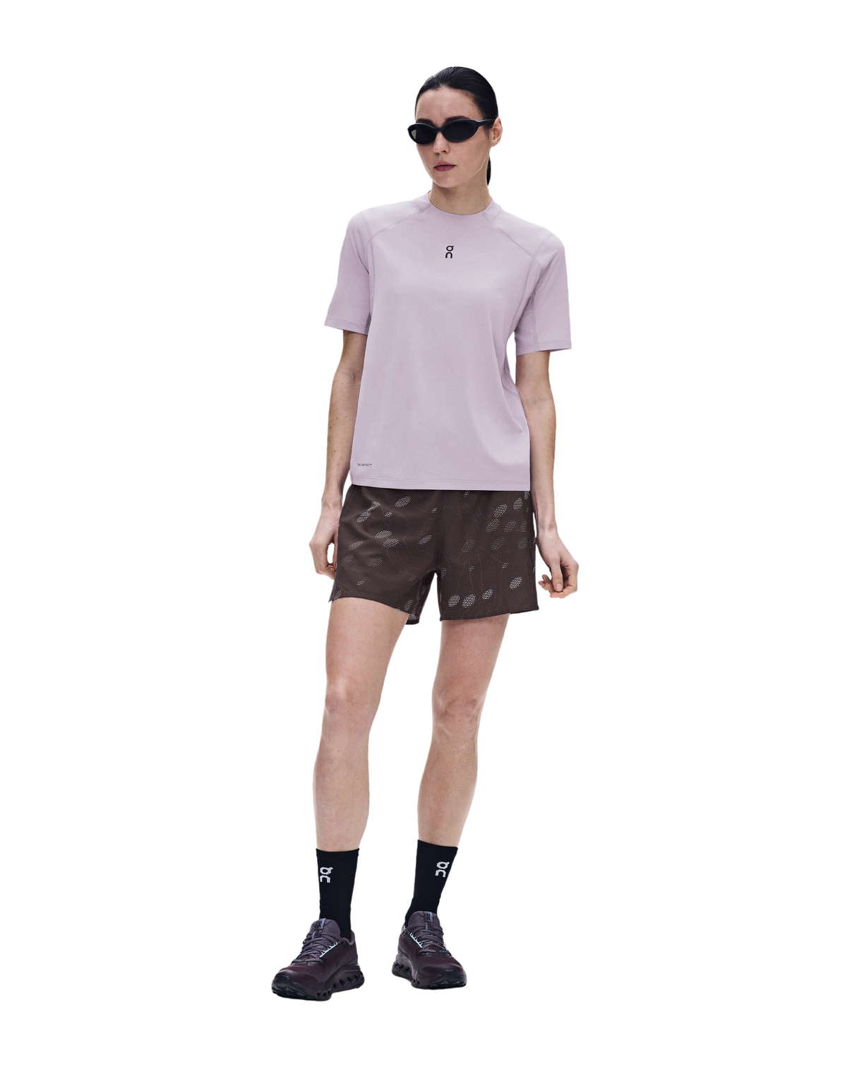 A model wearing a light purple athletic t-shirt, dark brown patterned shorts, black socks, and black running shoes, standing against a solid white background. - On Trail-T Solar (Women's) - Foxglove