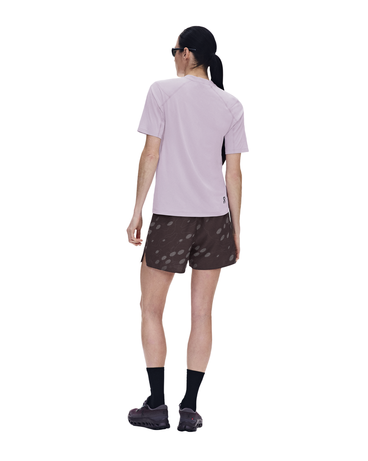 A model shown from the back wearing a light purple short-sleeve athletic shirt, brown patterned running shorts, black socks, and trail running shoes against a solid white background. - On Trail-T Solar (Women's) - Foxglove
