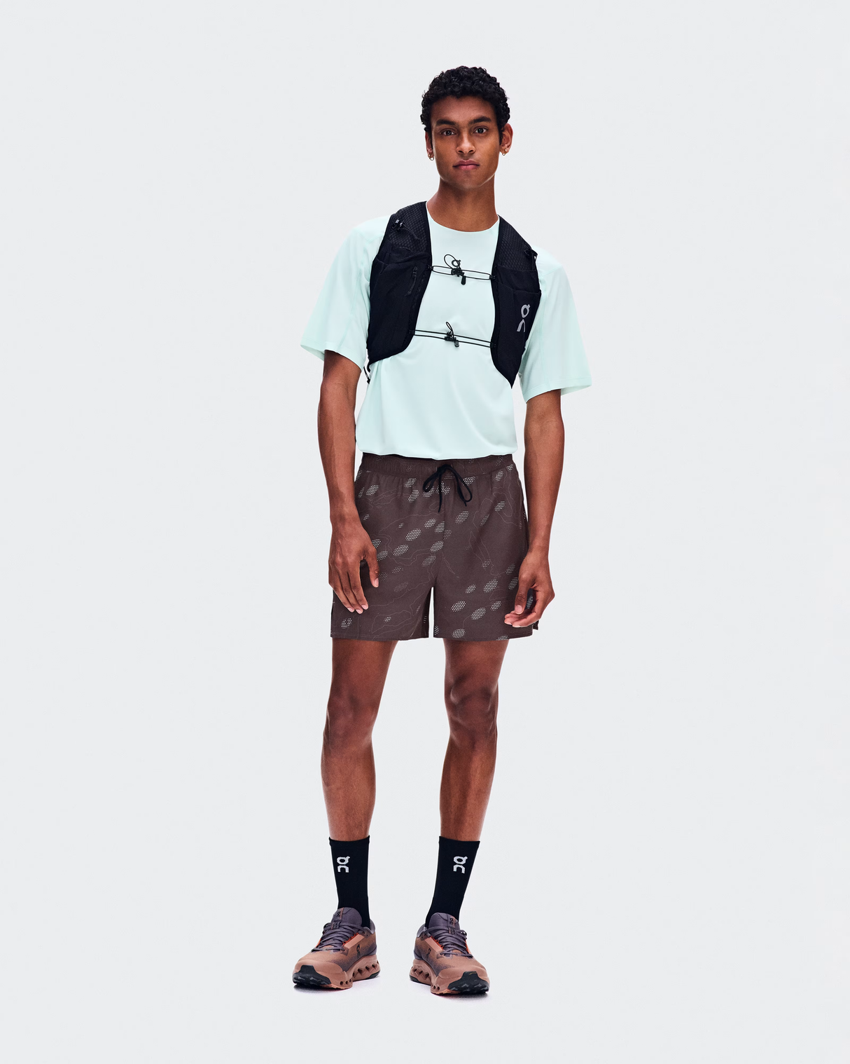 A male model stands against a light gray studio background wearing a white t-shirt, brown patterned shorts, black crew socks, trail running shoes, and a black hydration backpack. - On Trail Shorts (Men's) - Mulberry