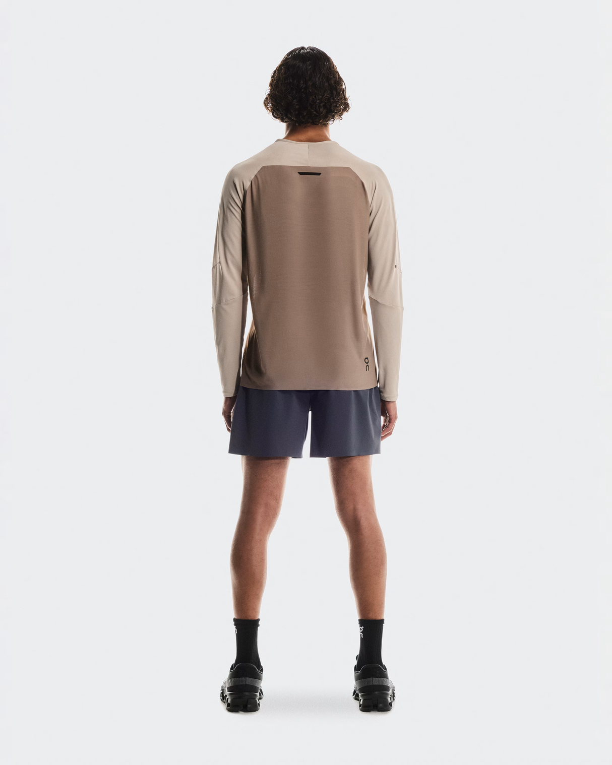 Person wearing a beige long-sleeve shirt and dark shorts on a white background - On Performance Long-T (Men's) - Desert/Cinder