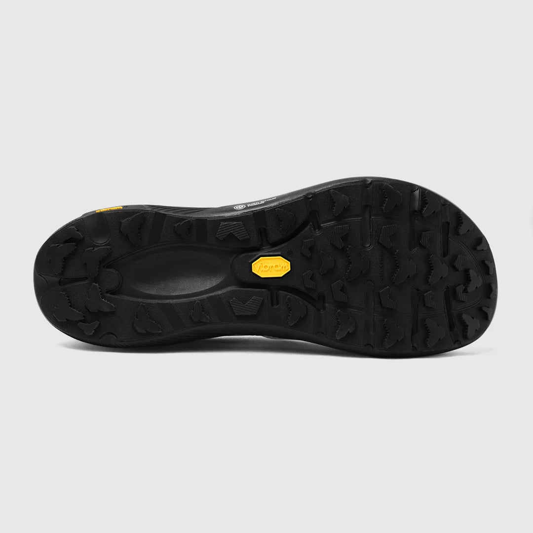 The black rubber outsole of a hiking shoe, featuring deep lugs and a small yellow Vibram logo badge in the center, shown against a light gray background. - Tarkine Trail Devil 3 Shoes (Men's) - Apex White