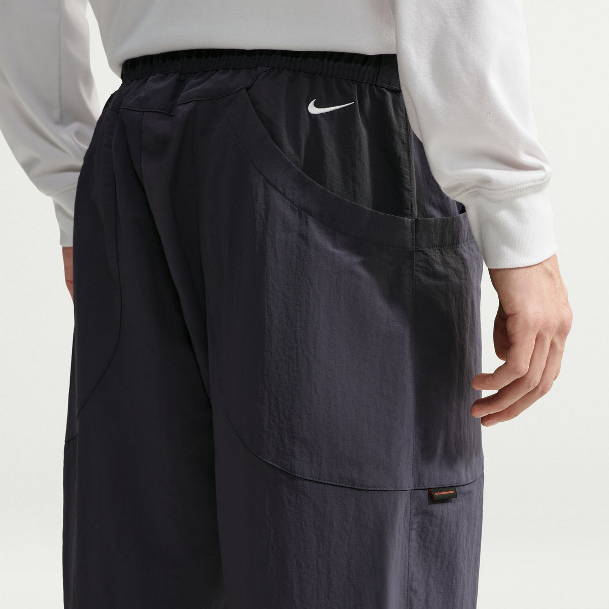 Close-up of a person wearing dark navy Nike pants with a white long-sleeve top, showing the back pocket and side logo detail. - Nike ACG Dolomiti Pants (Men's) - Gridiron/Summit White