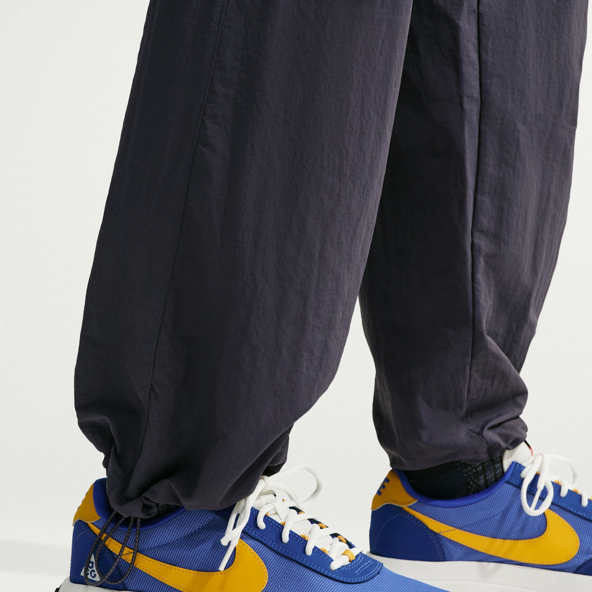 Close-up of a person's legs wearing dark charcoal gray jogger pants and blue and yellow Nike sneakers against a clean white background. - Nike ACG Dolomiti Pants (Men's) - Gridiron/Summit White