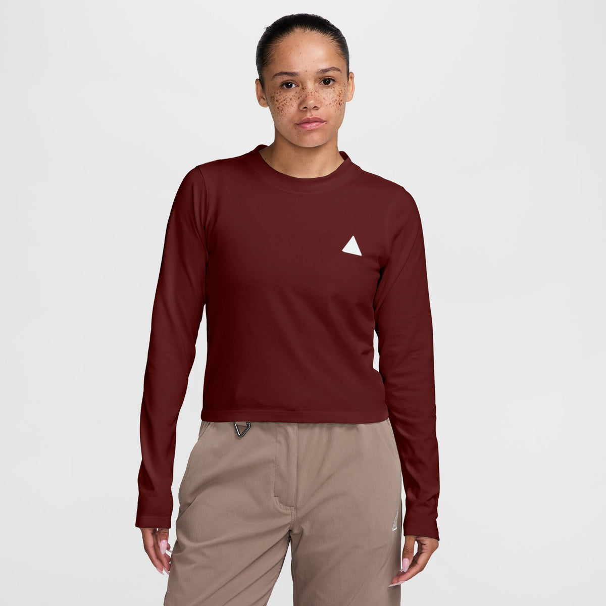 A woman modeling a long-sleeve crewneck shirt in deep burgundy with a small white geometric logo on the left chest, paired with beige pants, against a light gray studio background. - Nike ACG Dri-FIT Long-Sleeve Top (Women's) - Bold Berry/Summit White