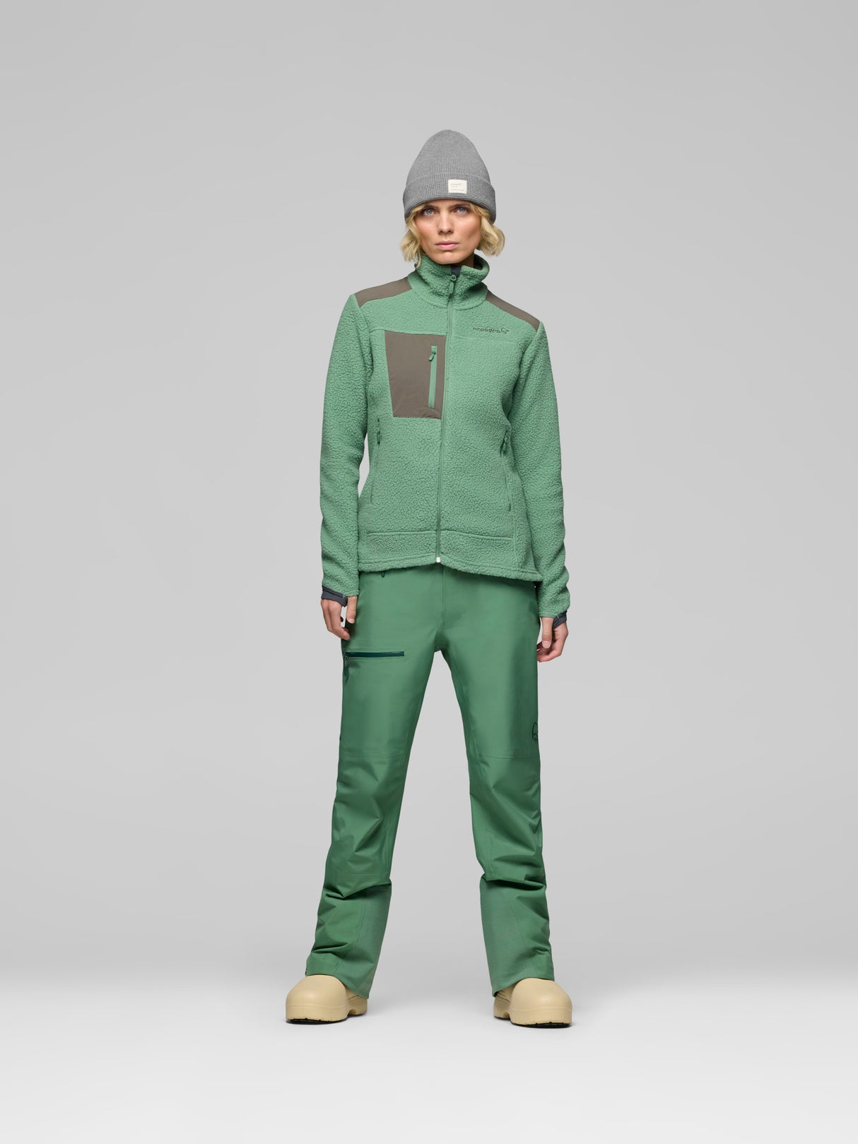 A model stands against a light gray studio background wearing a matching green outdoor fleece jacket and pants set, with a gray beanie and tan boots. - Norrøna Trollveggen Warm3 Jacket (Women's) - Dark Ivy