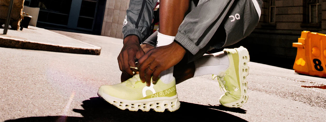 Person tying bright green ON CLOUDMONSTER 3 ROAD RUNNING SHOES on a street with a building in the background