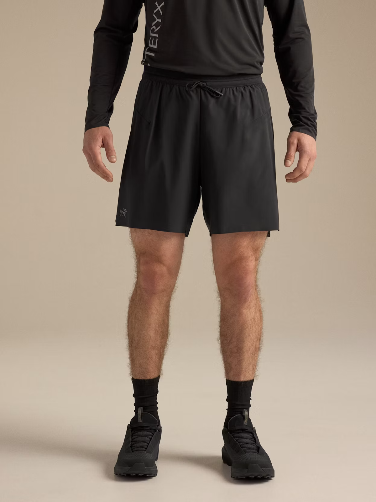 A model from the waist down wearing black athletic shorts, a black long-sleeve top, black socks, and black running shoes against a solid beige studio background. The shorts feature a drawstring waist and a small white logo on the left leg. - Arc'teryx Norvan Liner Shorts 7" (Men's) - Black