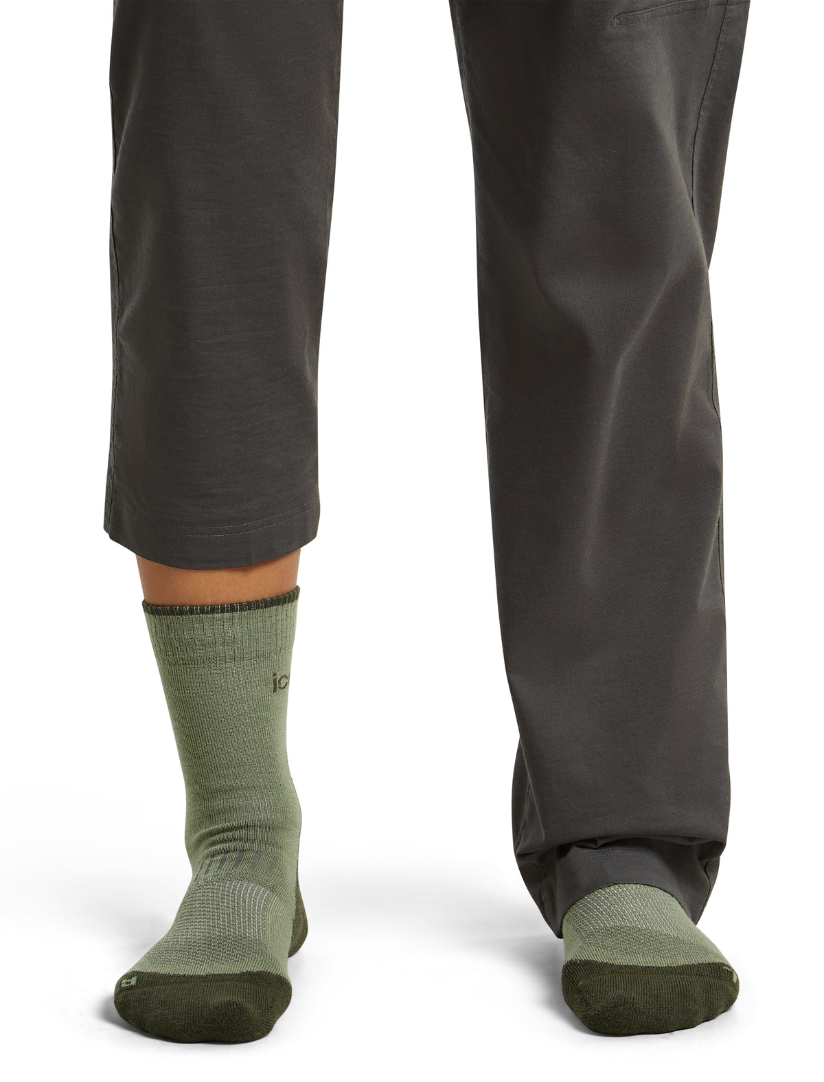 A close-up of a person's lower legs wearing dark gray pants and olive green crew socks with dark green toe and heel sections, shown against a plain white background. - Icebreaker Merino Hike+ Light Crew Socks (Women's) - Lichen/Dark Loden