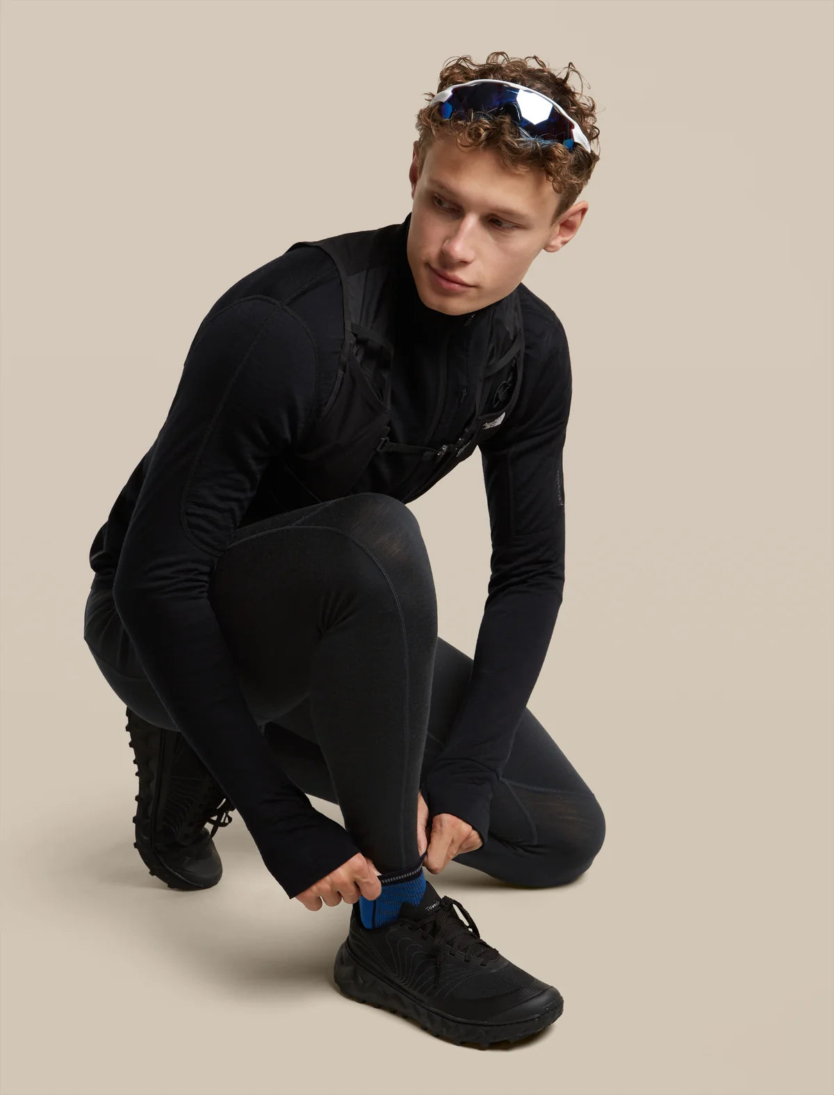 A man in a black long-sleeve athletic top and black running tights crouches while tying his black running shoe. He is wearing reflective sunglasses on his head and a blue sock on his ankle, set against a solid beige studio background. - Icebreaker Merino Run+ Ultralight Mini Socks (Men's) - Lazurite/Midnight Navy