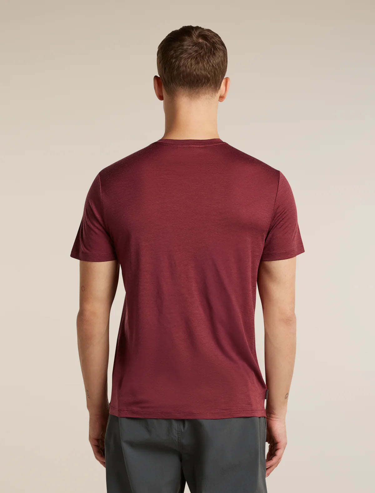 A man viewed from behind wearing a short-sleeve crew-neck t-shirt in a heathered burgundy red, paired with dark gray pants, against a plain beige studio background. - Icebreaker Merino 150 Tech Lite Short Sleeve T-Shirt (Men's) - Port