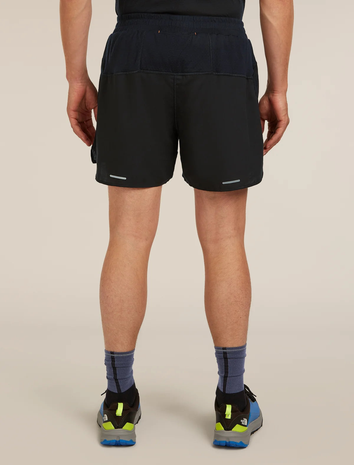 Rear view of a person wearing black athletic shorts, a dark navy top, blue socks, and running shoes against a plain beige studio background. - Icebreaker Merino Blend 125 ZoneKnit Speed 6" Shorts (Men's) - Black
