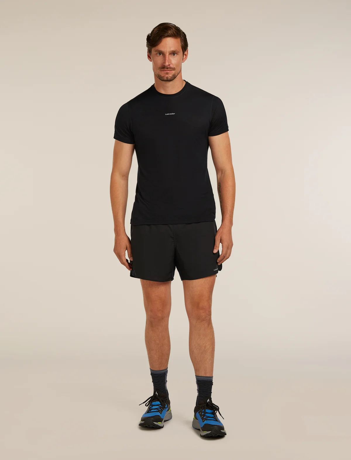 A man modeling a black short-sleeve athletic t-shirt and black running shorts, paired with dark socks and blue running shoes, against a plain beige studio background. - Icebreaker Merino Blend 125 ZoneKnit Speed 6" Shorts (Men's) - Black