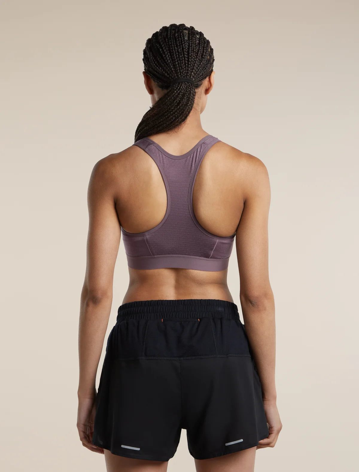 Rear view of a woman wearing a mauve-purple athletic sports bra and black athletic shorts against a plain beige studio background. - Icebreaker Merino Blend 125 ZoneKnit Racerback Bra (Women's) - Grey Plum