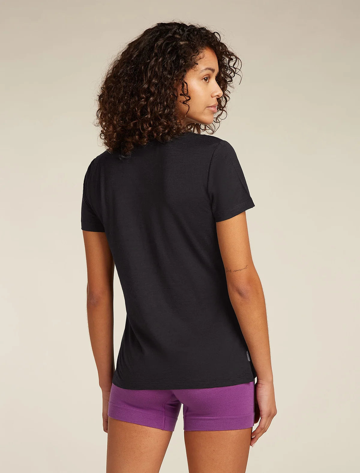 A woman with curly dark hair is shown from the back wearing a black short-sleeve t-shirt and purple athletic shorts against a plain beige studio background. - Icebreaker Merino 150 Tech Lite Short Sleeve T-Shirt Sparkling Stars (Women's) - Black