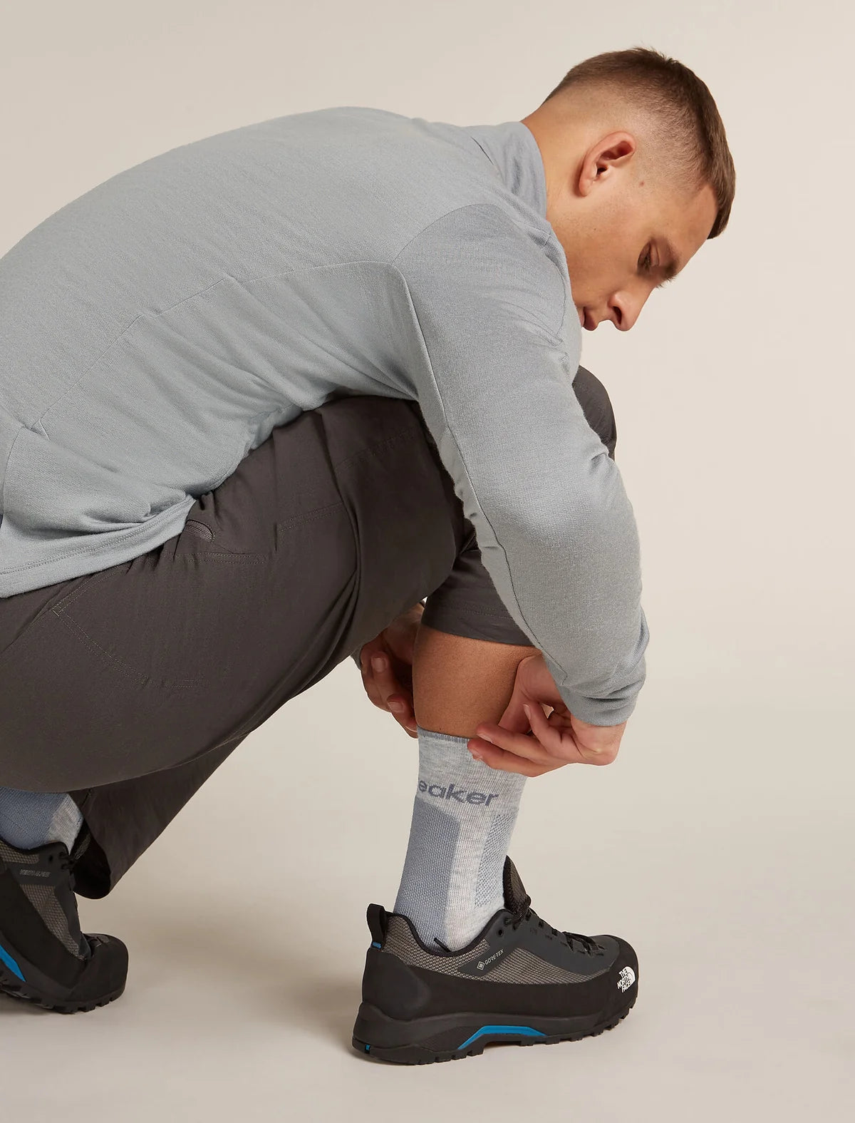 A man crouching down to show off a light gray crew sock with 'baker' printed on it, paired with a light gray long-sleeve shirt, dark gray shorts, and black athletic shoes with blue accents. - Icebreaker Merino Hike+ Medium Crew Socks (Men's) - Blizzard Heather/Alloy