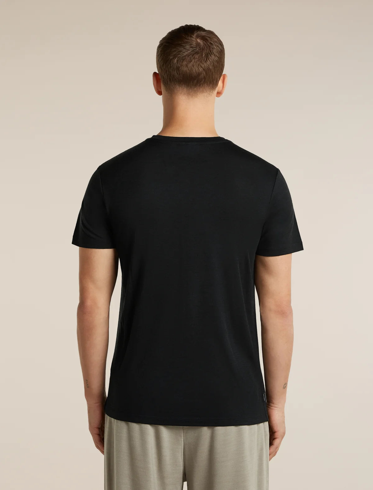 A man viewed from the back wearing a plain black short-sleeve crew-neck t-shirt and light beige trousers, standing against a solid light beige background. - Icebreaker Merino 150 Tech Lite Short Sleeve T-Shirt Range Stripes (Men's) - Black