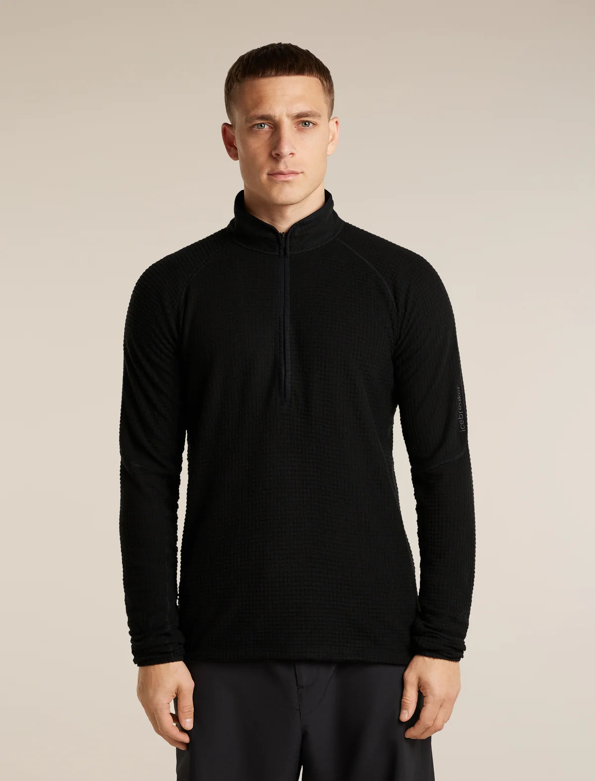 A man wearing a black long-sleeve quarter-zip athletic pullover with a textured waffle knit pattern, standing against a solid light beige background. - Icebreaker Merino 400 RealFleece Grid Peaksland Long Sleeve Half Zip (Men's) - Black