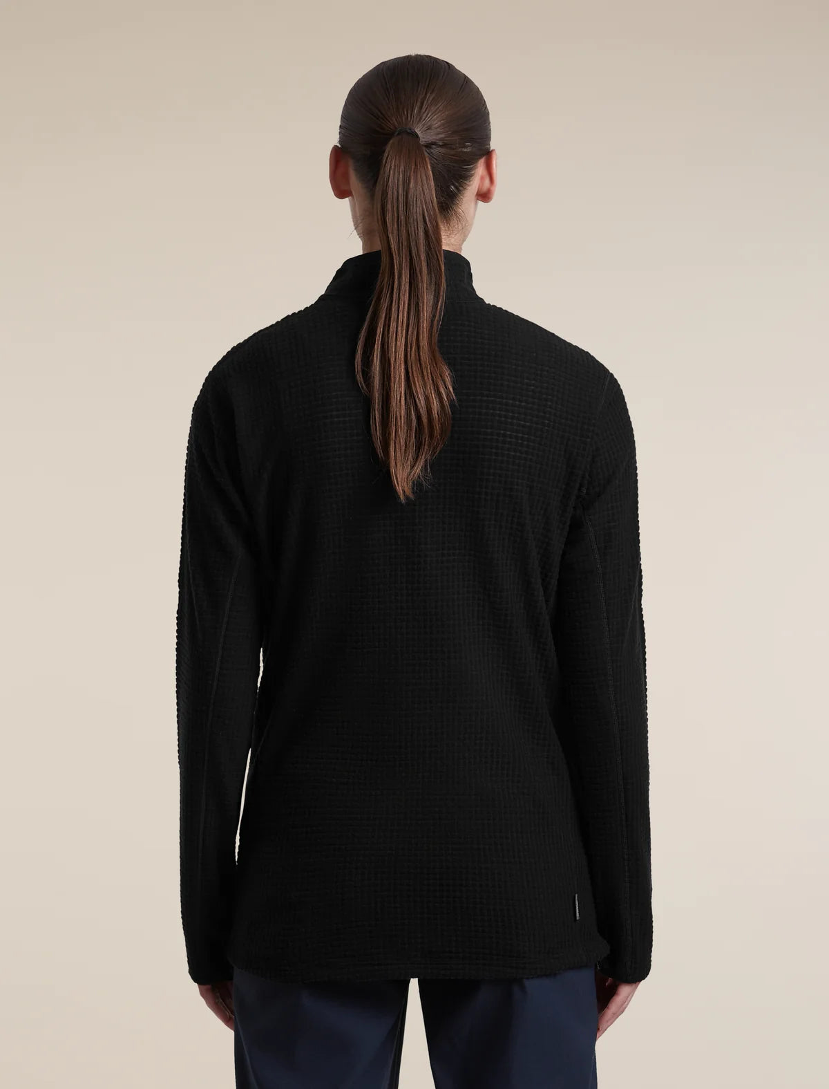 A woman viewed from the back wearing a black textured quarter-zip pullover sweater and dark trousers, set against a plain beige studio background. - Icebreaker Merino 400 RealFleece Grid Peaksland Long Sleeve Half Zip (Women's) - Black