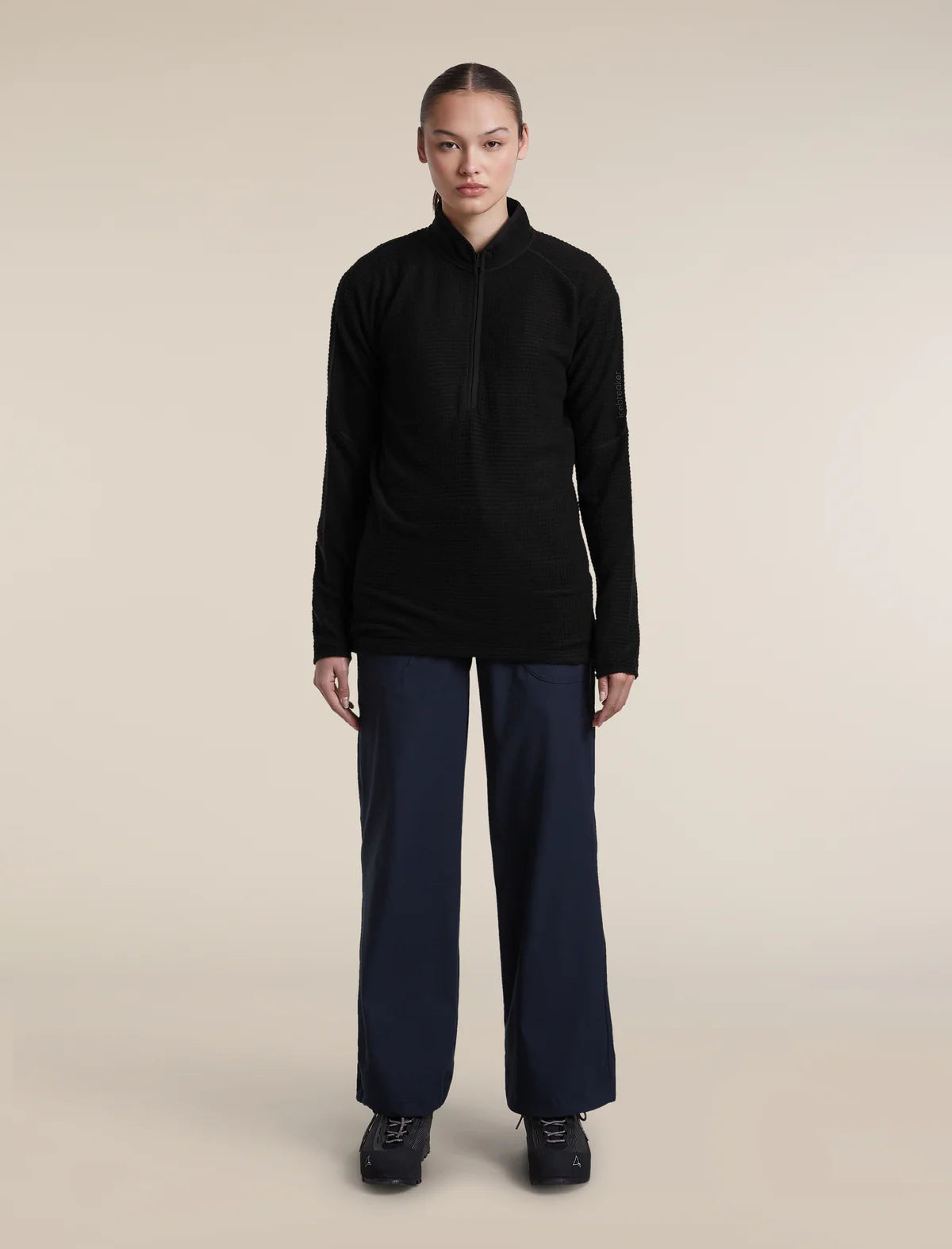 A model stands against a plain beige studio background wearing a black long-sleeve half-zip pullover, dark navy wide-leg trousers, and black shoes. - Icebreaker Merino 400 RealFleece Grid Peaksland Long Sleeve Half Zip (Women's) - Black