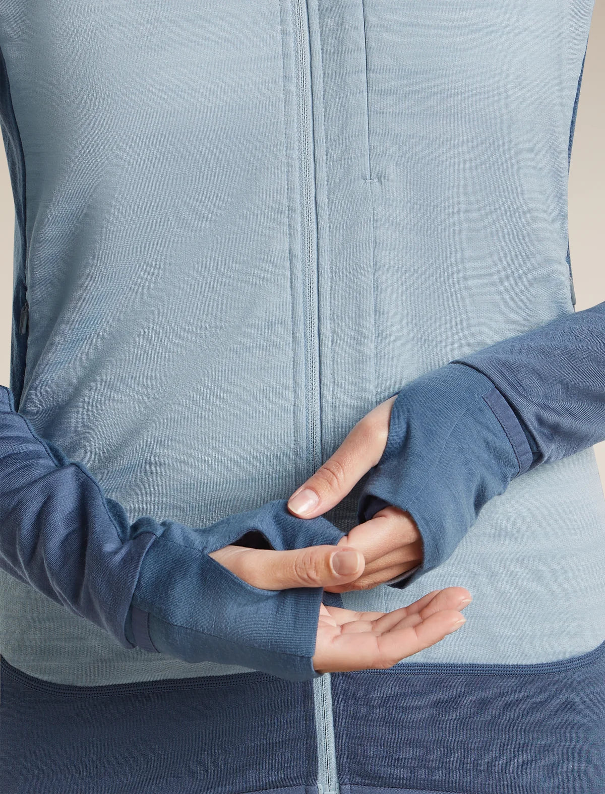 Close-up of a person wearing a blue long-sleeve athletic zip jacket, showing the cuffs and torso against a plain light beige studio background. - Icebreaker Merino Blend 300 RealFleece Descender Long Sleeve Zip Hoodie (Women's) - blue, light blue, dawn/ zinc blue