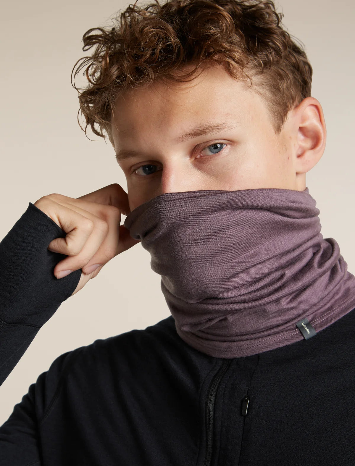 A close-up of a man wearing a dark plum purple neck gaiter pulled up over his nose and mouth, paired with a black long-sleeve top against a solid beige background. - Icebreaker Merino 200 Flexi Chute (Unisex) - Grey Plum