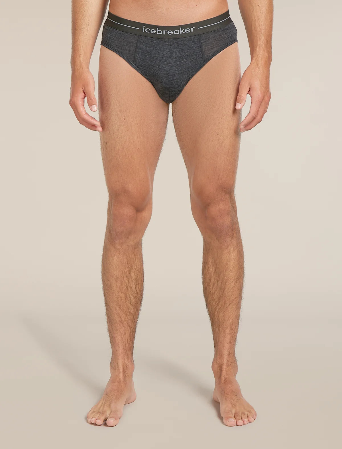 A man wearing dark gray heathered briefs with a black waistband featuring the white 'icebreaker' logo, shown from the waist down against a light beige studio background. - Icebreaker Merino 150 Anatomica Briefs (Men's) - Jet Heather