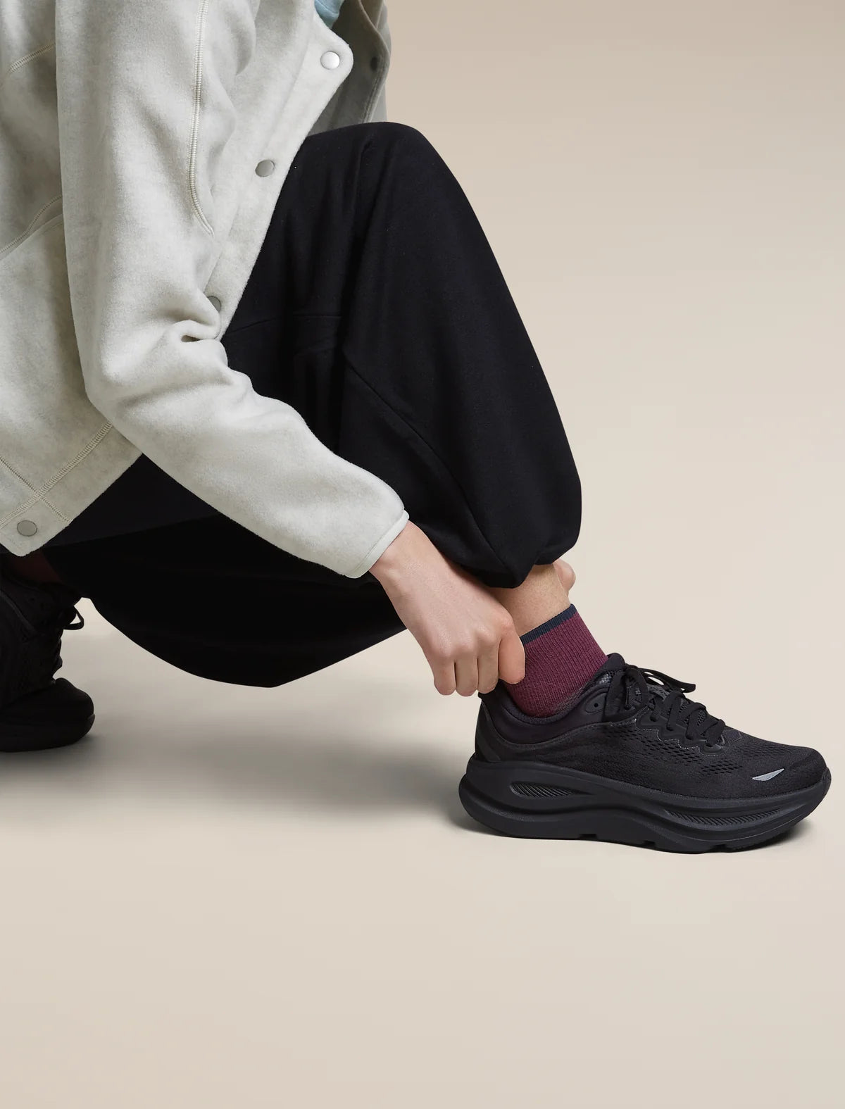 A person crouching and pulling up a black athletic sneaker with a chunky sole, wearing black pants, a light gray cardigan, and a burgundy sock, against a plain beige studio background. - Icebreaker Merino Multisport Light Mini Socks (Women's) - Dark Berry/Midnight Navy/Grey Plum