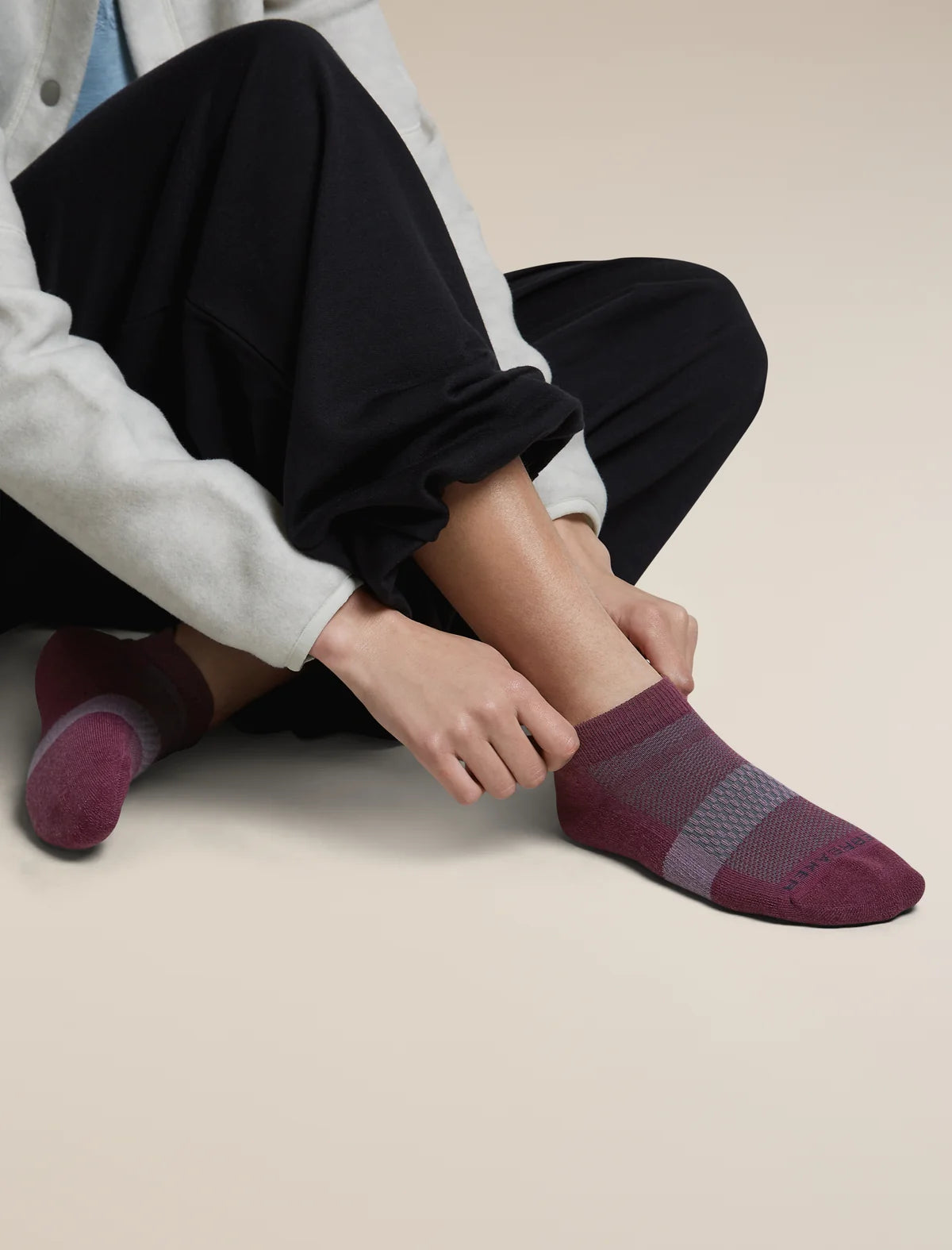 A person sitting on a light beige floor wearing dark pants and light gray sleeves, adjusting a low-cut knit sock on one foot. The sock is burgundy with a gray knit stripe across the instep. - Icebreaker Merino Multisport Light Micro Socks (Women's) - Dark Berry/Midnight Navy/Grey Plum