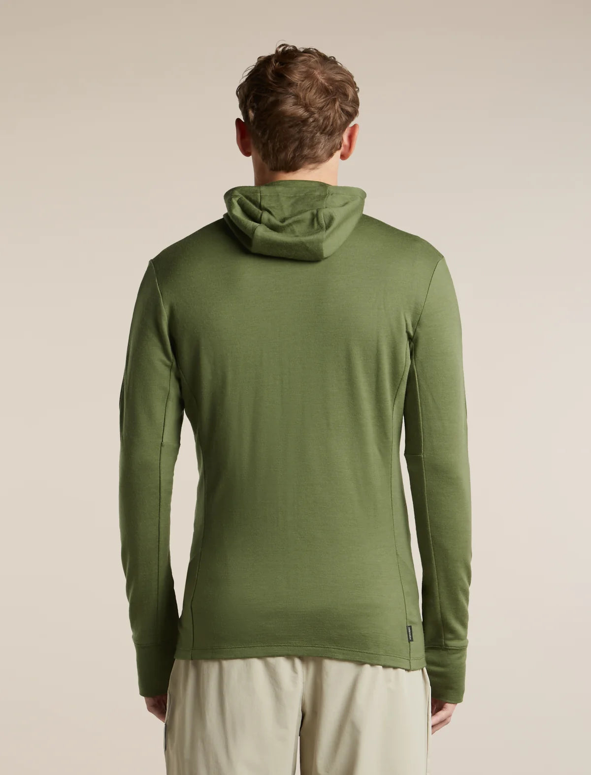 A man viewed from behind wearing a sage green hooded long-sleeve shirt and light beige pants, set against a plain beige studio background. - Icebreaker Merino 260 Quantum Long Sleeve Zip Hoodie (Men's) - Sphagnum