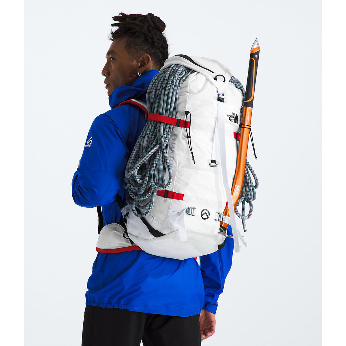A person wearing a bright blue jacket carries a large white technical backpack with red straps, gray climbing ropes, and an orange-and-black ice axe against a light gray studio background. - The North Face Phantom 38 Backpack - TNF White/Raw Undyed-NPF