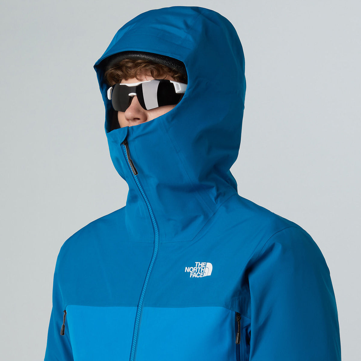 Person wearing a blue hooded jacket with a brand logo on a gray background - The North Face Jazzi 3L GORE-TEX Jacket (Men's) - Skyline Blue/Adriatic Blue
