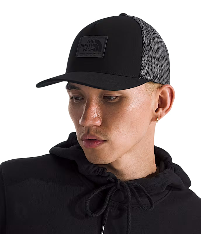 A man wearing a black trucker hat with a black mesh back and a black patch logo for The North Face, paired with a black hoodie against a white background. - The North Face Keep It Patched Trucker - TNF Black/Asphalt Grey