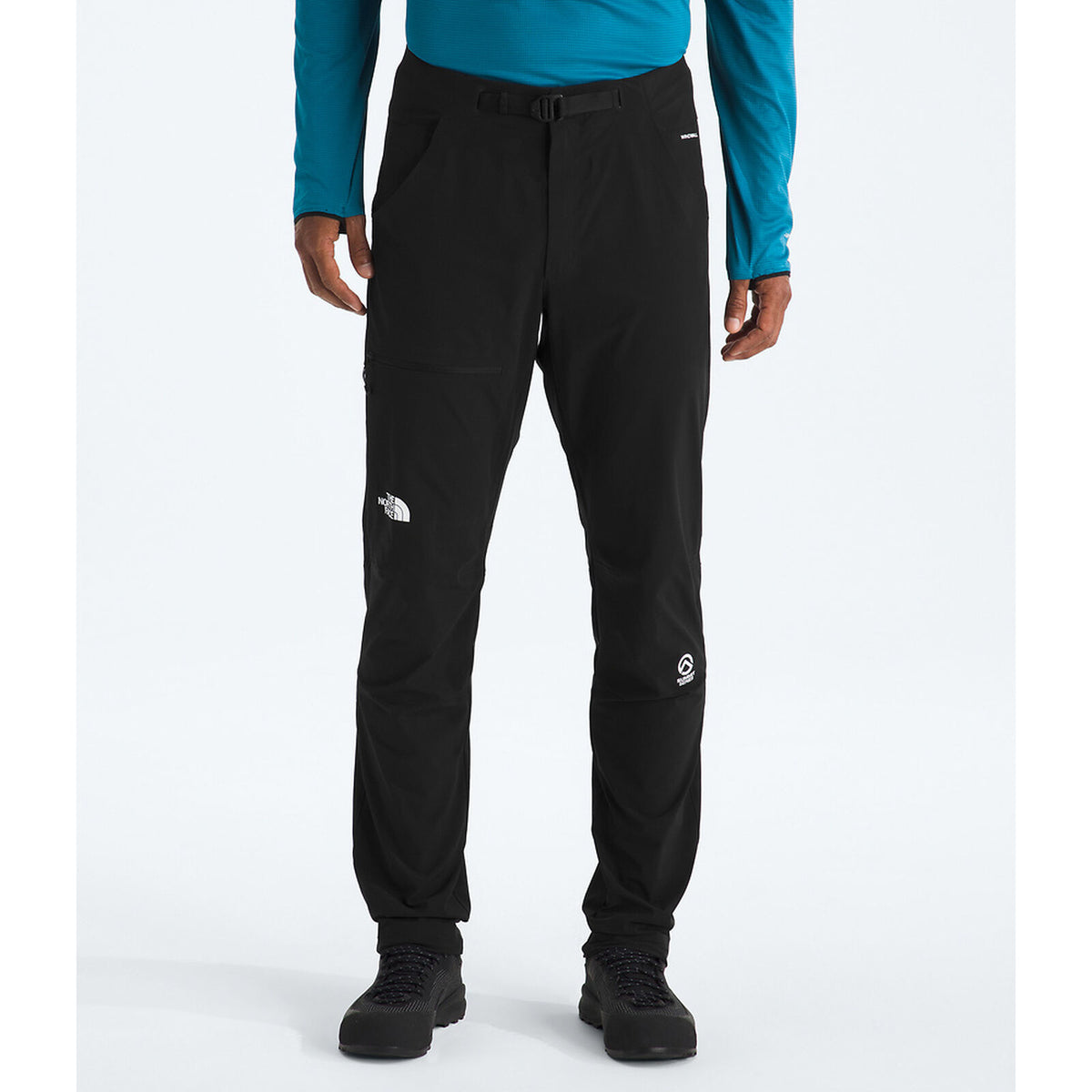 A person wearing black technical pants with white logo patches, a teal long-sleeve top, and black shoes, photographed against a clean light gray studio background. - The North Face Summit Summit Series Off Width Pants (Men's) - TNF Black