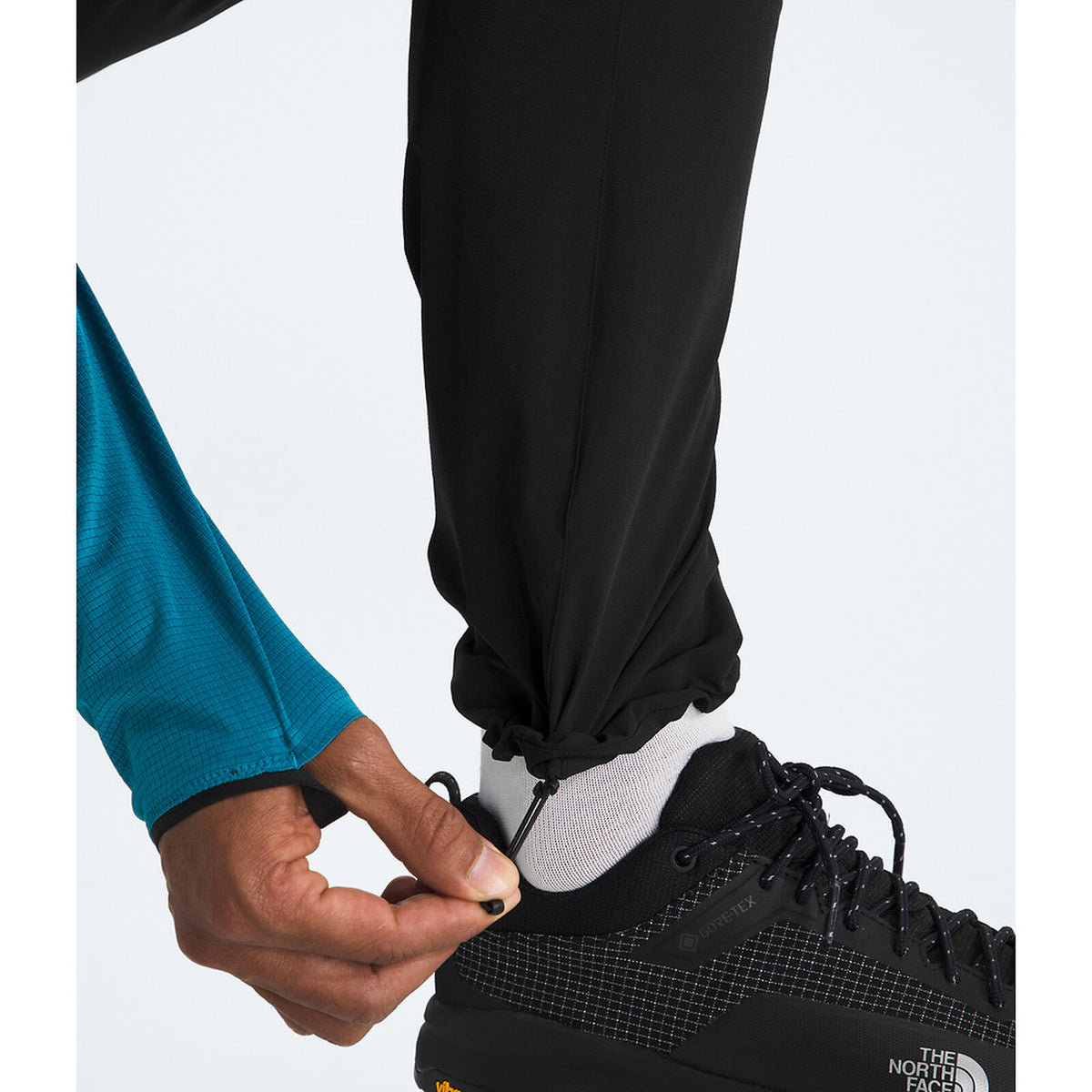 A close-up of a person's leg and hand adjusting the elastic cuff of black pants, wearing a white sock and a black The North Face running shoe. - The North Face Summit Summit Series Off Width Pants (Men's) - TNF Black