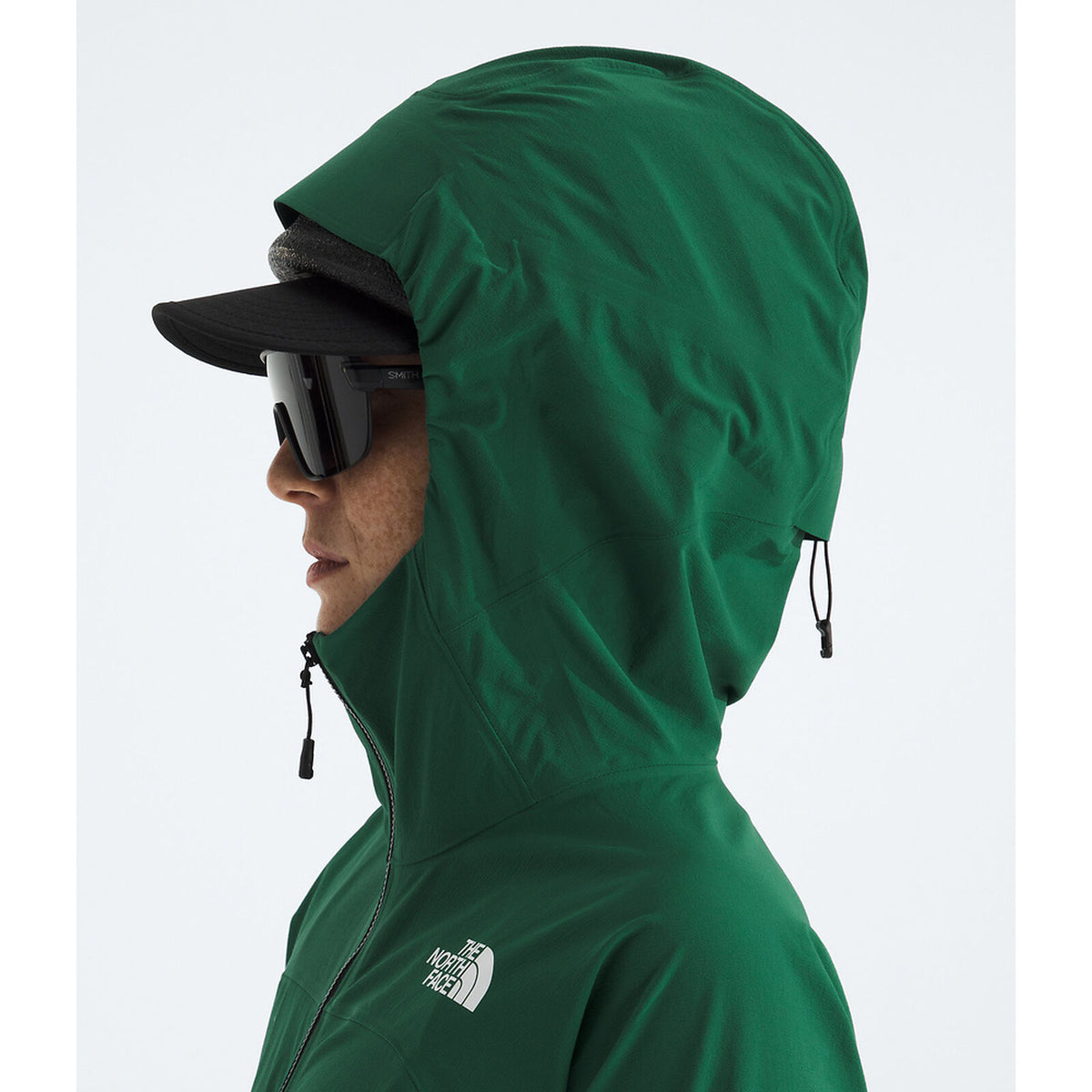 A side profile of a person wearing a green hooded jacket by The North Face, a black cap, and dark sunglasses, against a light gray background. - The North Face Summit Series Off Width Jacket (Women's) - Evergreen