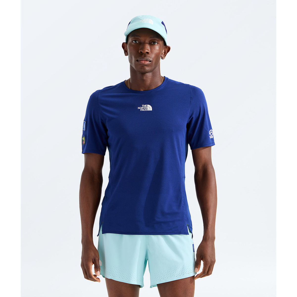 A male model wearing a royal blue short-sleeve athletic t-shirt by The North Face, paired with light blue shorts and a matching light blue cap, set against a plain light gray studio background. - The North Face Summit High Trail Short Sleeve-Graphic (Men's) - Bold Blue