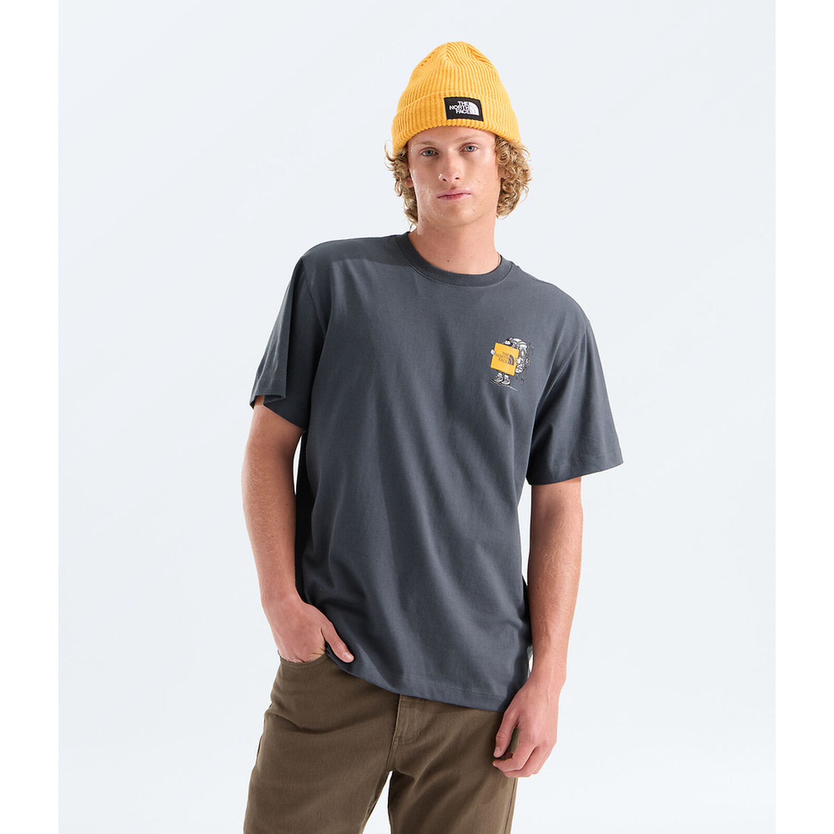 A man wearing a dark charcoal gray t-shirt with a small yellow graphic on the left chest, paired with a yellow beanie and brown pants, standing against a light gray studio background. - The North Face Gear Up Get Out Relaxed Short-Sleeve Tee-Graphic (Unisex) - Anthracite Grey