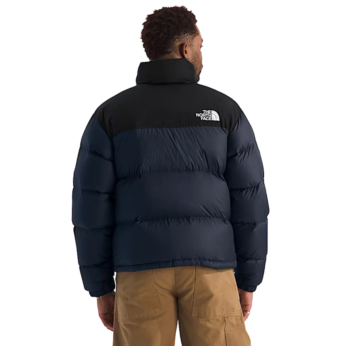 A man viewed from the back wearing a puffer jacket with a black upper section and a dark navy lower section, paired with tan cargo pants against a plain white background. - The North Face 1996 Retro Nuptse Jacket (Men's) - Summit Navy/TNF Black