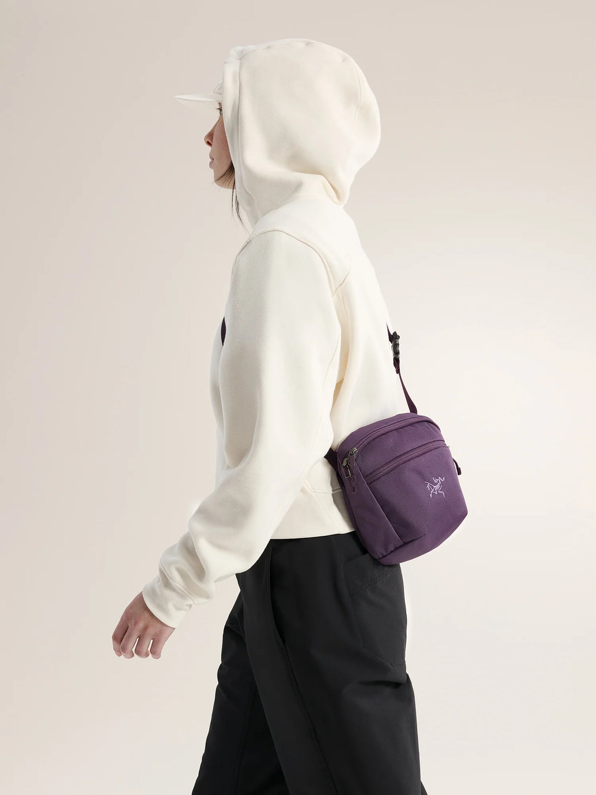 A person in a white hoodie and black pants shown in side profile, wearing a small purple crossbody bag with a visible Arc'teryx logo on the front pocket, against a plain light beige studio background. - Arc'teryx Mantis 2 Waist Pack - Moondrop