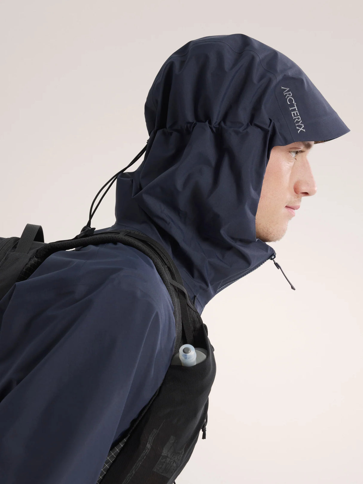 A man in profile wearing a dark navy hooded rain jacket and a black backpack, shown against a plain light background. The Arc'teryx logo is visible on the hood. - Arc'teryx Beta Jacket (Men's) - Black Sapphire