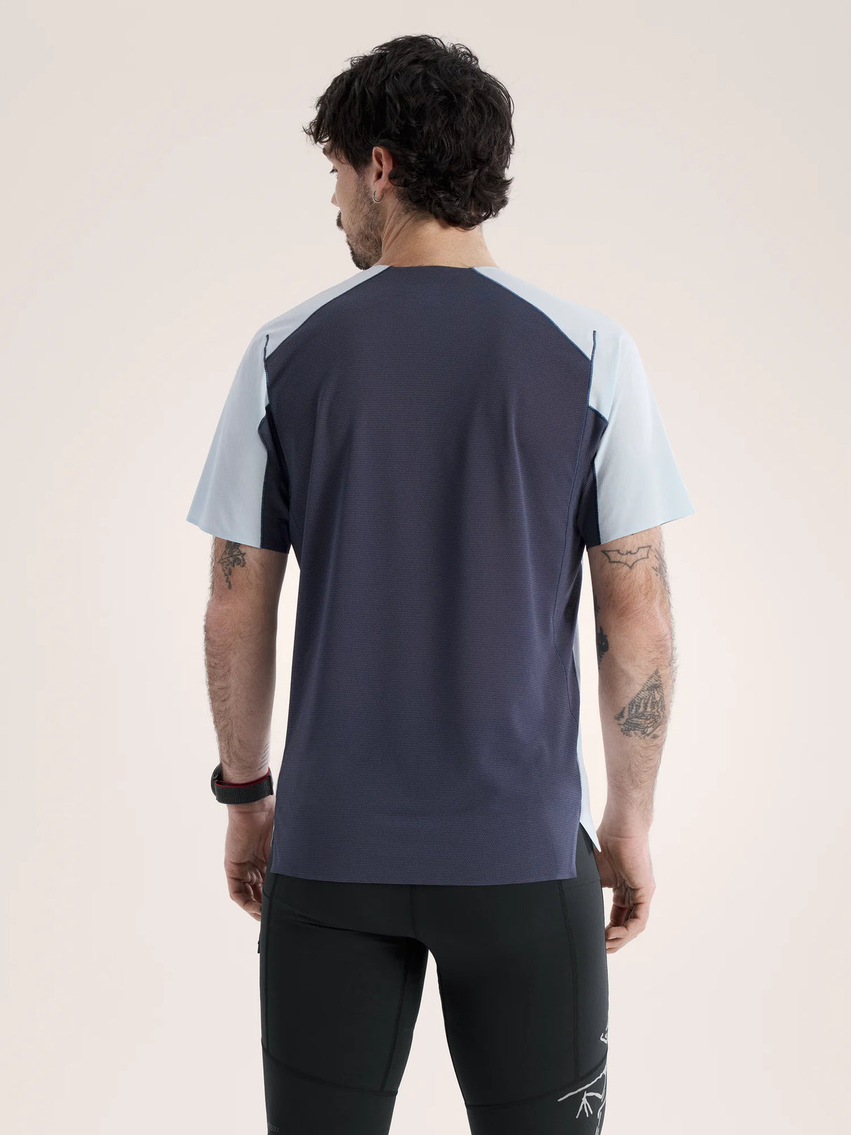 A man viewed from the back wearing a short-sleeve athletic shirt with a dark navy body and light blue shoulder panels, paired with black athletic pants, against a plain light beige studio background. - Arc'teryx Norvan Downword Logo Shirt Short Sleeve (Men's) - Alpine Blue/Dark Stratus