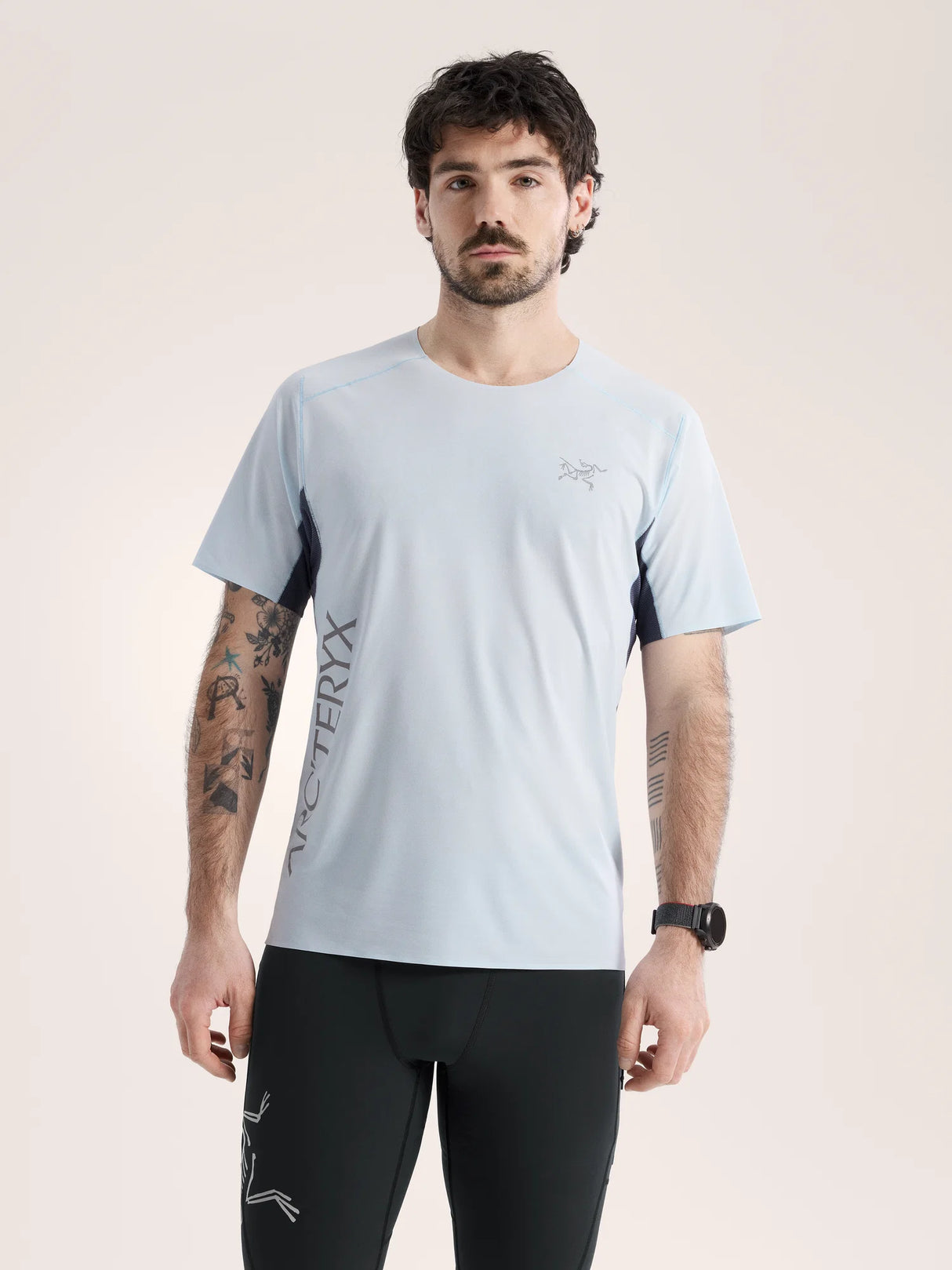 Man wearing a light blue athletic shirt with black sleeves and black pants on a beige background - Arc'teryx Norvan Downword Logo Shirt Short Sleeve (Men's) - Alpine Blue/Dark Stratus