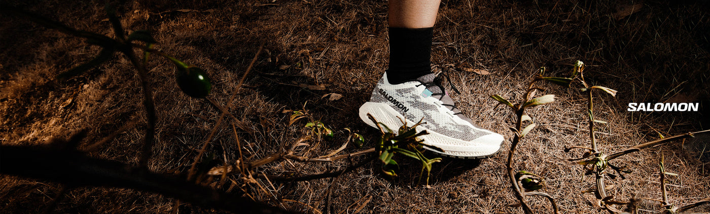 Person wearing white SALOMON AERO GLIDE 4 GRVL with black socks on a dark, natural ground surface, featuring the Salomon brand.