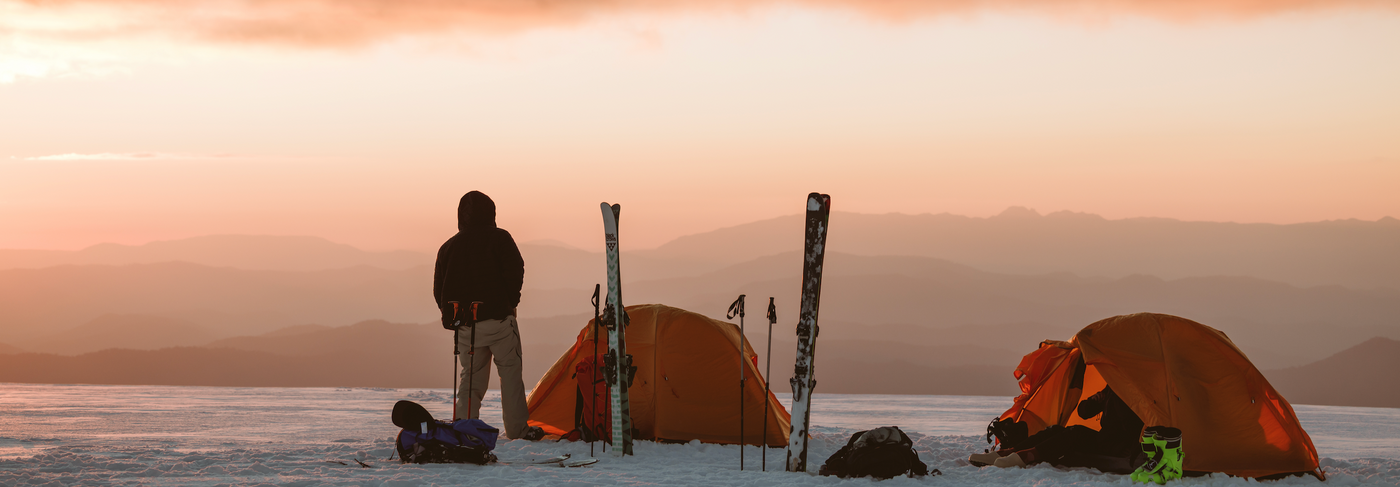Person with skis and two orange One Planet tents on a snowy landscape at sunset.