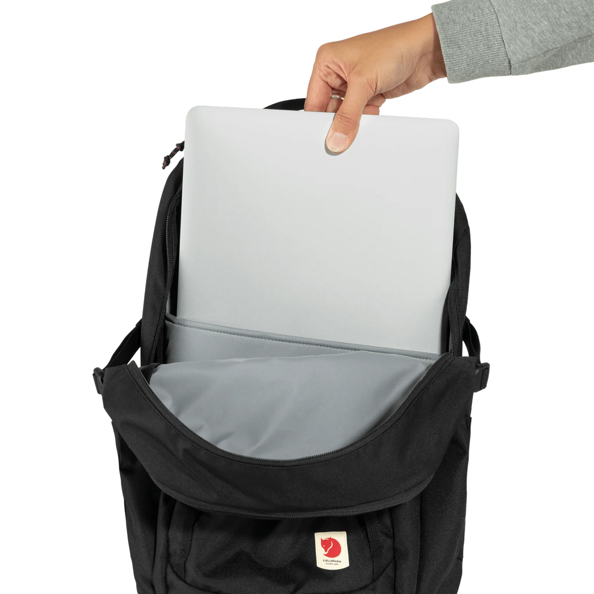 A black Fjälör laptop backpack shown open with a hand inserting a silver laptop into the top compartment. The backpack features a gray interior lining and a small red and white Fjälör logo patch on the front pocket, set against a plain white background. - Fjällräven Skule 28 Backpack - Black