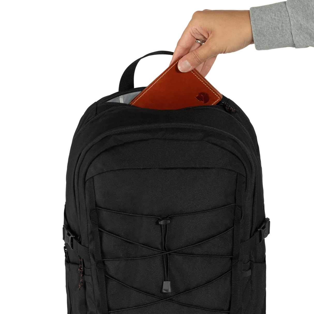 A black backpack shown from the front with a hand inserting a brown leather wallet into the top zipper compartment, against a plain white background. - Fjällräven Skule 28 Backpack - Black