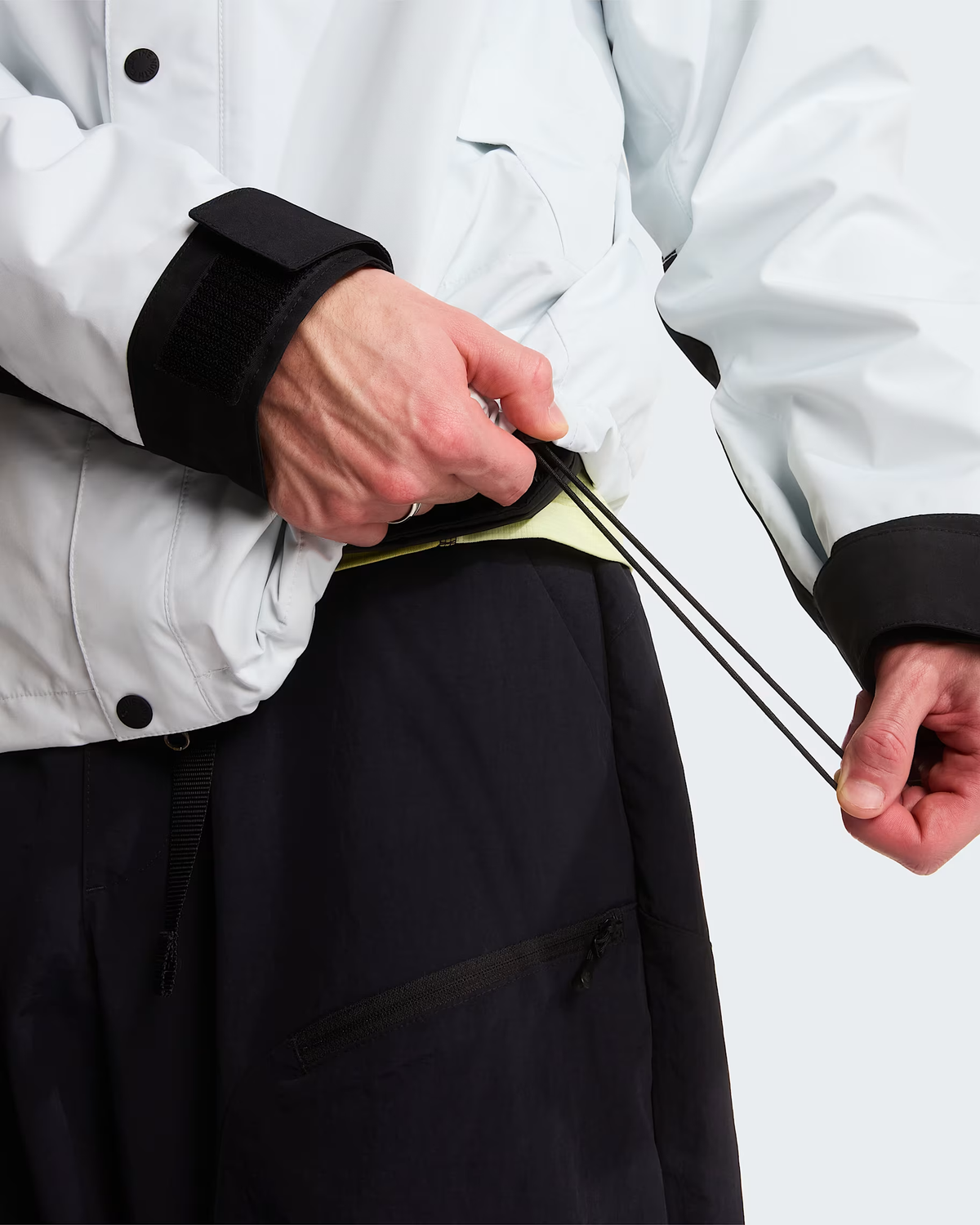 Close-up of a person wearing a light gray jacket with black cuffs and dark pants, pulling a black drawstring at the waist. - The North Face GORE-TEX Mountain Jacket (Men's) - Pearl Stone/TNF Black